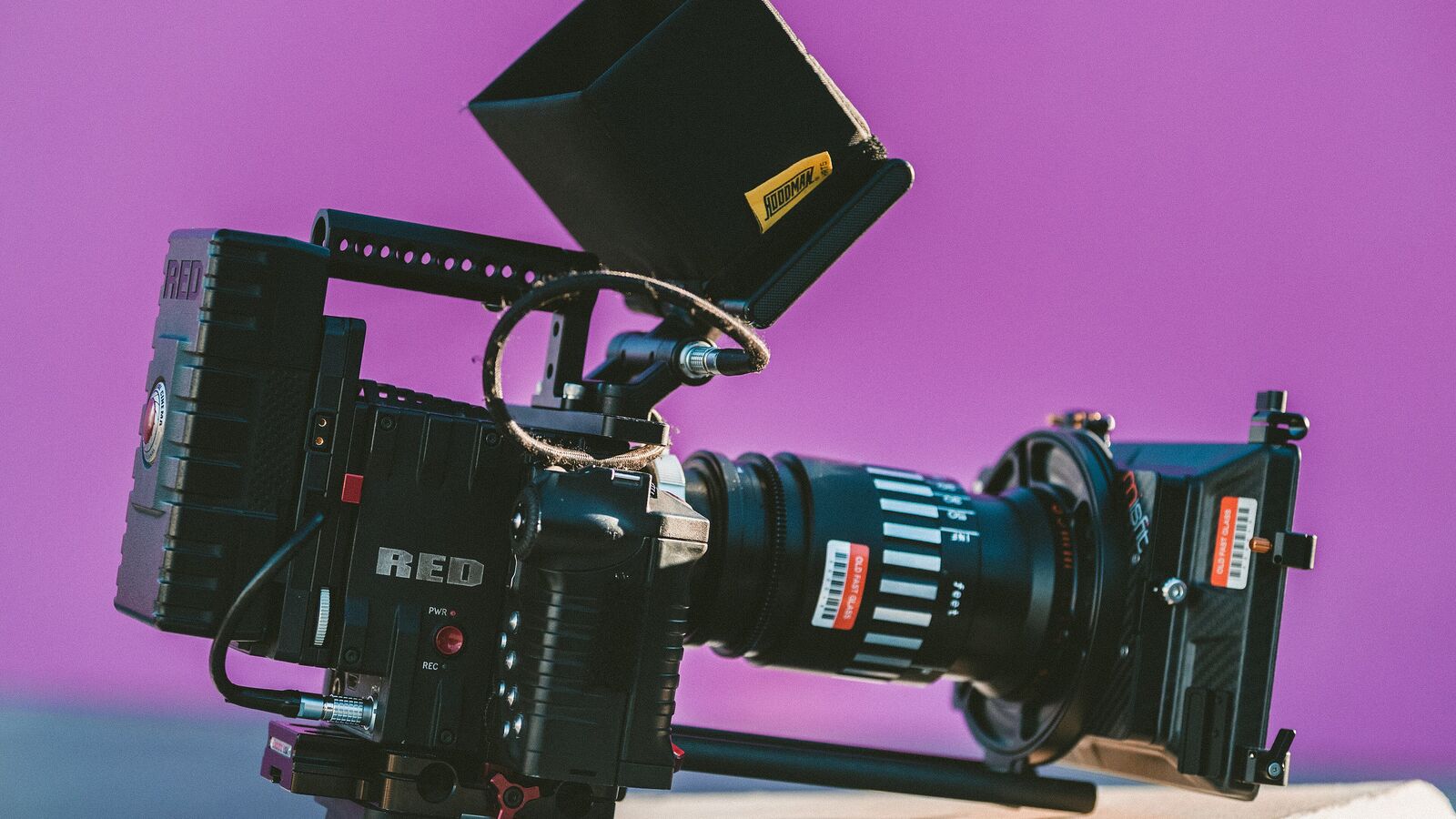 A black cinema camera with a matte box and lens in front of a purple backdrop
