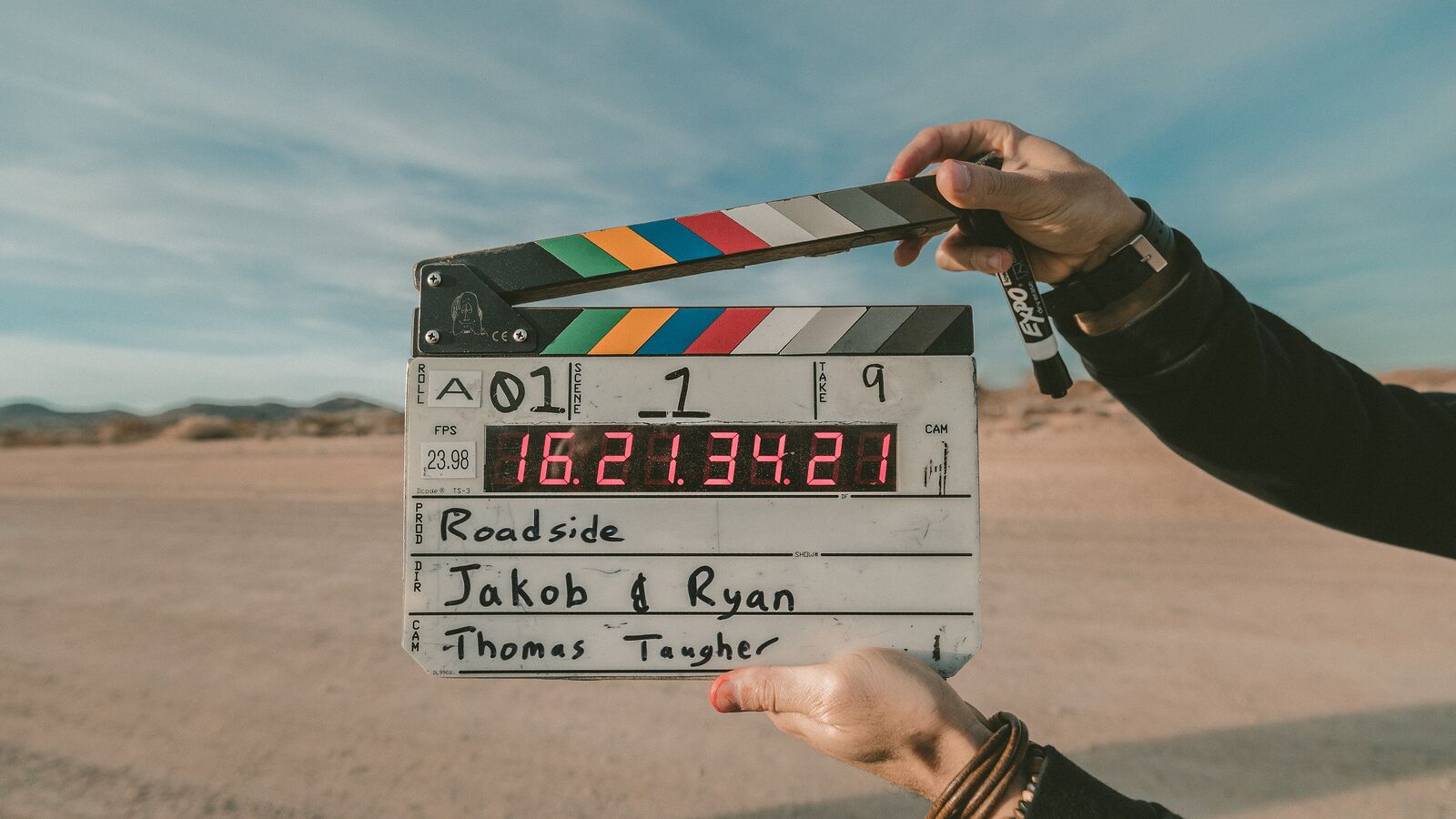 Hands holding an open clapperboard in front of a sandy landscape and sky