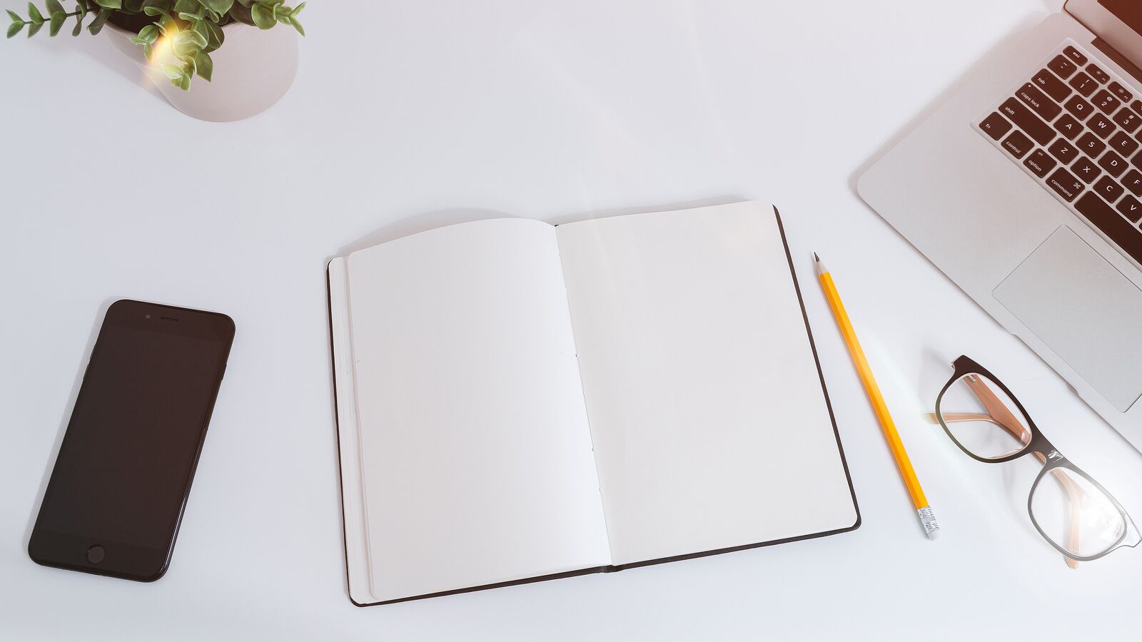 Top view of a white desk with an open blank notebook, a pencil, glasses and a smartphone