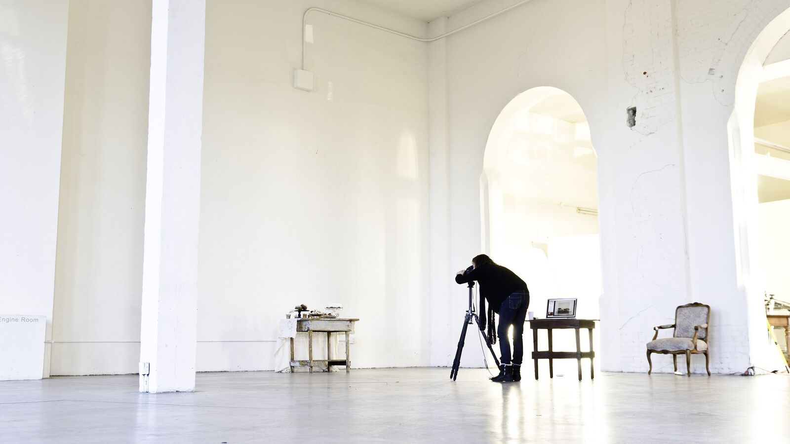 A large, empty white room with tall arches and a distant person at a tripod