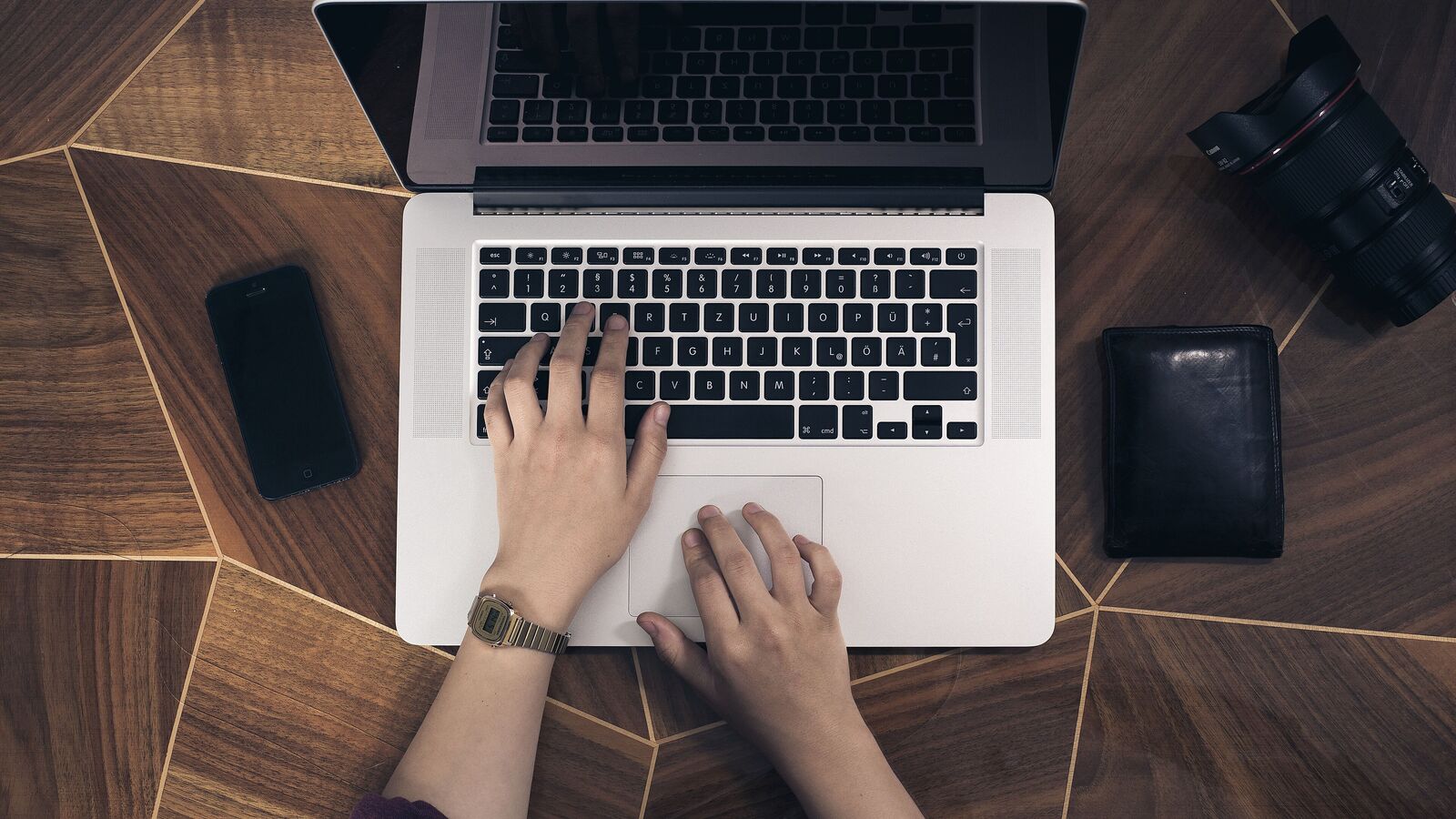 Top view of two hands typing on a laptop on a wooden desk, with a smartphone and a camera lens beside it