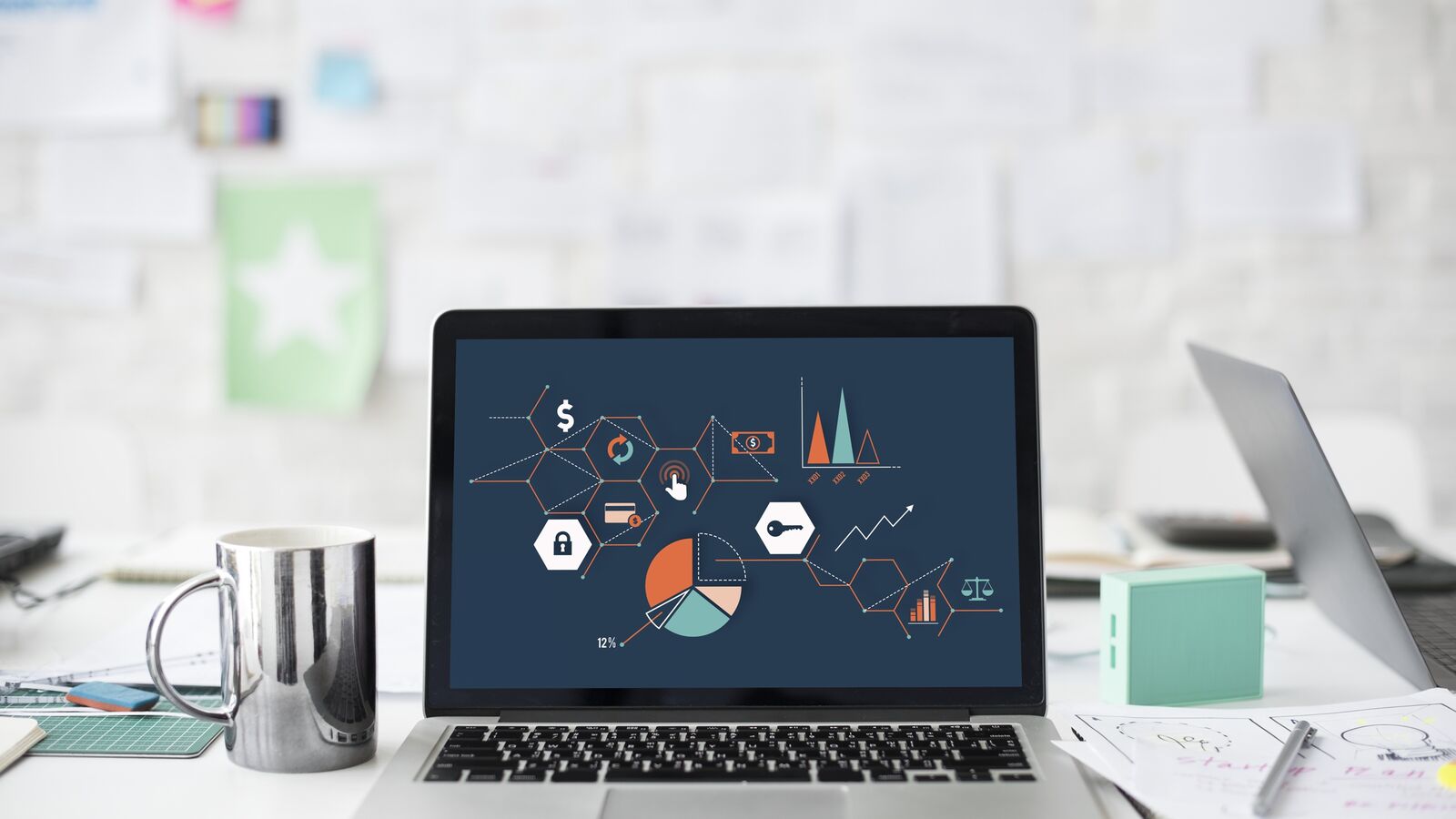 A laptop on a bright desk showing a dashboard with charts, next to a metal mug
